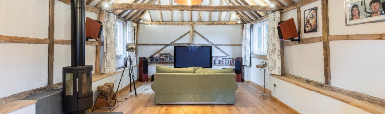 Spacious attic lounge with exposed timber beams, a wood-burning stove, and a comfortable seating area focused around a large television.